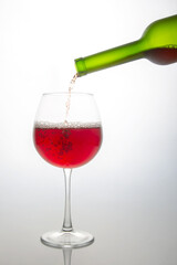 A wine bottle pouring rich red wine into a clear glass goblet, capturing the moment of pour with bubbles forming. Set against a soft white background, perfect for wine tasting or celebration themes