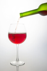 wine bottle pouring rich red wine into a clear glass goblet, capturing the moment of pour with bubbles forming. Set against a soft white background, perfect for wine tasting or celebration themes