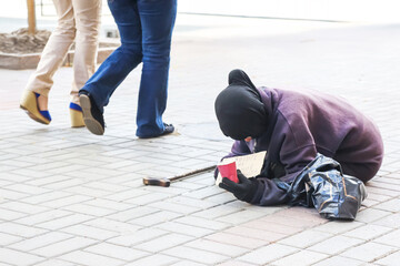 A person in a purple hoodie and black headscarf sits on a pavement, holding a sign and a cup,...