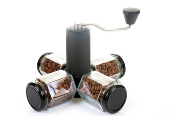 black manual coffee grinder for coffee beans and four glass transparent jars with black lids filled with roasted coffee beans and earthenware for brewing coffee on a white background.