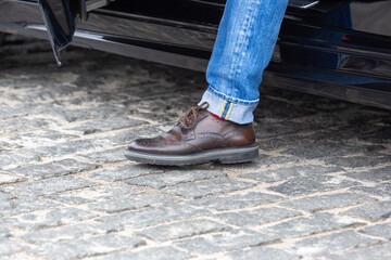 A person in blue jeans and brown brogue shoes steps out of a black car onto a cobblestone street,...
