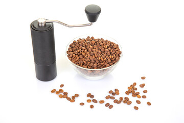 modern manual coffee grinder next to a transparent bowl overflowing with rich brown coffee beans, with a few beans spilled on a pristine white surface