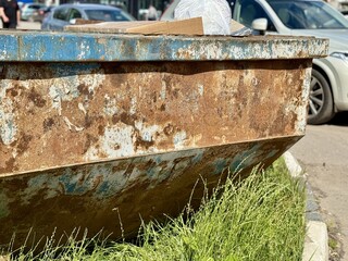 An Old and Rusty Dump Container That is Surrounded by Lush, Overgrown Grass and Weeds. A weathered metal dumpster overflowing with various debris, positioned against a backdrop of lush tall grass.