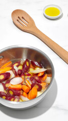 Vegetables being prepared in a silver bowl with wooden spoon and olive oil on a kitchen counter.
