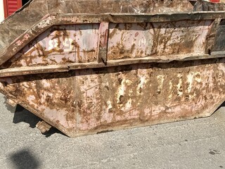 An old rusty dumpster sitting on a bustling city street filled with urban life and debris. The dumpster resting on the pavement clearly shows numerous signs of urban decay and waste management issues.