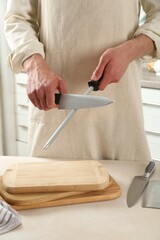 Man sharpening knife with sharpener at beige table indoors, closeup