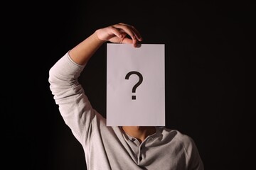 Man hiding behind paper sheet with question mark on dark background