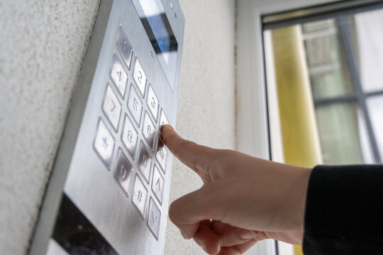 A person is pressing a button on a sleek security entry system attached to a wall in an urban setting while sunlight filters through nearby windows