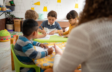 Kindergarten students color pictures at a classroom table with the guidance of their preschool...