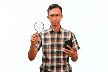 An Asian man holding a magnifying glass and a mobile phone with a serious expression