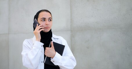 Professional businesswoman wearing hijab standing near gray wall while making phone call, holding...