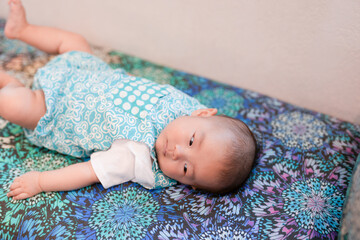 A Japanese baby celebrating their 100th day lies on a colorful sofa, surrounded by cushions. Their...