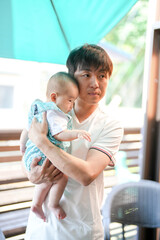 A Japanese father in his thirties holds his 100-day-old baby on a sunlit terrace with potted...