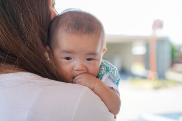 In bright sunshine, a Japanese mother in her twenties stands in her home's driveway garage, holding her 100-day-old baby close and cradling the child with soft, gentle warmth under a clear blue sky.