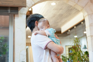 Fototapeta premium In a bright living room, a Japanese father in his thirties sits on a red sofa cradling his 100-day-old baby close amid soft cushions, the child's eyes wide with wonder as warm light fills the space.