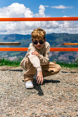 Latino boy crouching with confident gaze in sunny mountain setting.