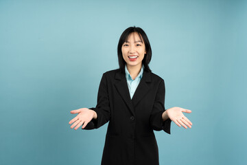 Smiling Asian woman in a black blazer gestures warmly against a blue background while engaging in...