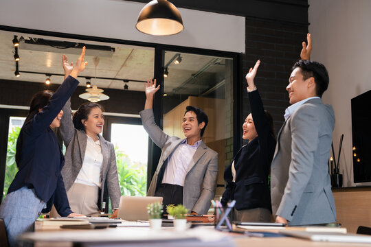 Team of happy diverse company employees raising hands for good agreement in corporate meeting. Group of motivated young and mature mixed race business people celebrating unity and cooperation.
