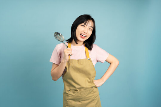 Young Asian woman in an apron holding a cooking utensil with a cheerful expression in a studio setting with a blue backdrop