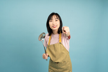 A joyful Asian woman in a casual pink shirt and yellow apron stirs a pot with a ladle, creating a...