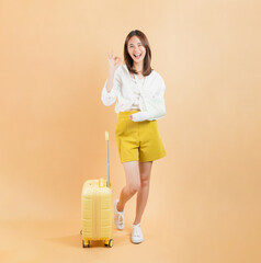 Asian woman broken arm wrapped and shows ok sign with suitcase isolated on beige color background, Travel insurance concepts.