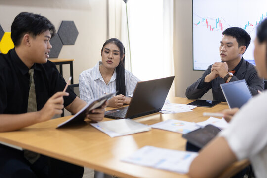 A group of young professionals engages in a dynamic meeting at a modern office, focusing on project strategies and collaboration while reviewing data on laptops.