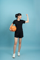 Confident Asian woman stands tall with a basketball in one hand, flexing her arm to showcase strength. She wears a black athletic outfit, embodying determination and sportsmanship.