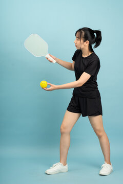 Asian girl practicing pickleball indoors with a paddle and yellow ball, showcasing athleticism and focus in casual sportswear