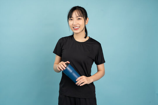 Young Asian woman in athletic attire holding a blue gym bottle against a solid blue background while smiling happily after a workout session