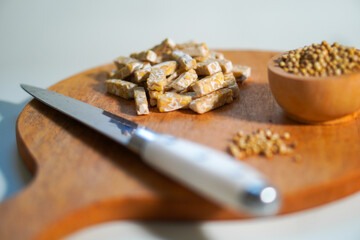 Tempeh that has been cut into small pieces on a wooden chopping board, complete with a knife and a bowl of coriander