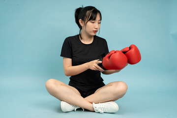 Young woman in black outfit sits cross-legged holding red boxing gloves against a blue background...