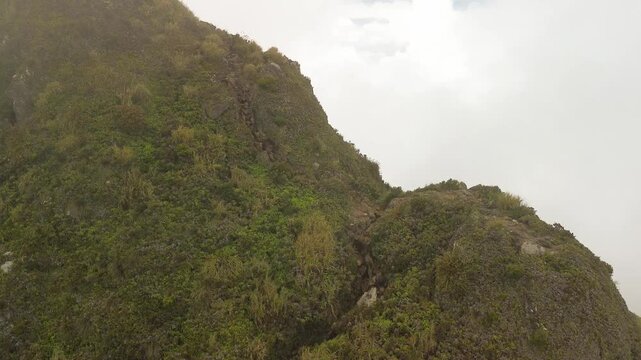 Steep volcanic slopes clothed in alpine grass and moss rise beside the lens, exposing a jagged ridge that pierces pale cloud layers high on Mount Apo.