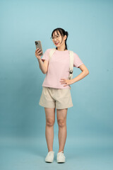 Young Asian woman standing with a smartphone in hand while smiling against a light blue background
