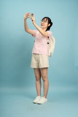 Young Asian woman takes selfie in a studio with light blue background, wearing casual attire with a bag and smiling happily