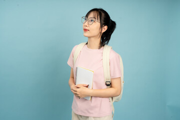 Young Asian student stands against a blue background with a backpack and notebook, contemplating...