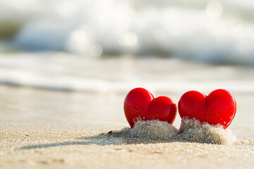 Fototapeta premium A close-up captures two shiny red heart-shaped objects resting on wet sand, with foamy ocean waves gently washing ashore in the background.