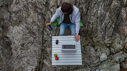 View from above. A white man sits at a table with a laptop on a cliff and holds a remote control...