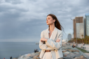 confident business woman in light suit standing with crossed arms near sea shore with city skyline...