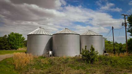 Rural scene featuring large grain silos under a vibrant blue sky, reflecting agricultural industry. Power lines and a farmyard add detail to this picturesque, working farm environment.