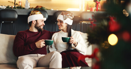 Christmas, happy and couple with coffee in home for festive morning with santa hat for celebration. Smile, love and man with woman on sofa for relax, bonding and drinking eggnog for xmas together.