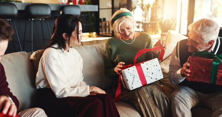 Christmas, gift and old woman with family on sofa for holiday season, bonding and support. Festive celebration, smile and xmas present with people in living room of home for love, package and relax