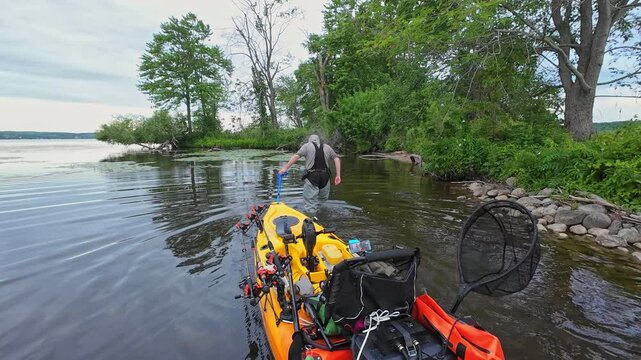 Angler pulling his kayak or water craft to the lake shore for brake, during fishing pov. Fishing pedal kayak in calm lake waters and peaceful landscapes. Water sports bass and pike fishing.