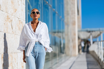 Obraz premium Fashion blogger standing in sunlight, slim asymmetric jeans and white shirt half-tucked in, 