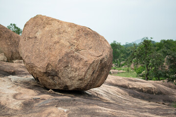 A massive boulder rests precariously on a rocky slope, surrounded by lush greenery and distant hills under a cloudy sky, showcasing nature's delicate yet powerful balance. © Bhudev