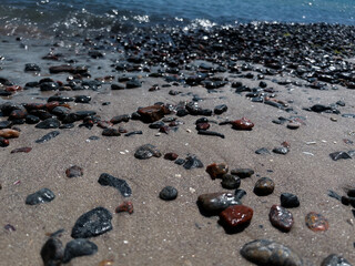 Close-up of wet pebbles scattered on a sandy beach in Odesa, Ukraine, with gentle sea waves sparkling in the background. The sunlit stones create a natural texture and coastal atmosphere, perfect for
