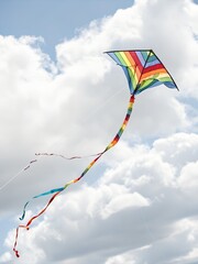A vibrant rainbow kite with a long colorful tail soars high in a cloudy blue sky