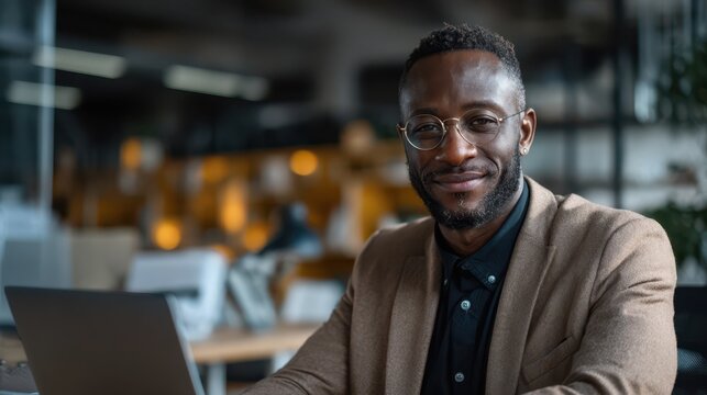 Confident young businessman wearing glasses and a smart blazer smiling at his desk in a modern open-plan office, representing professional success, leadership, and positive workplace culture