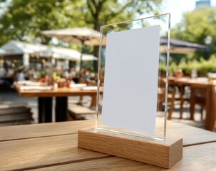 A blank menu or sign holder stands on a wooden table at an outdoor cafe with blurred chairs, tables, and greenery in the background.