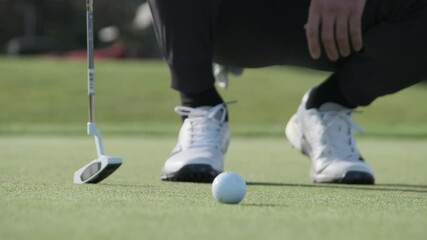 Man carefully lines up putt near golf ball on green during sunny day - Powered by Adobe