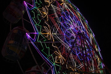 Colorful neon lights on a spinning Ferris wheel captured at night. Motion blur creates a vibrant pattern, symbolizing carnival fun and nighttime amusement rides.